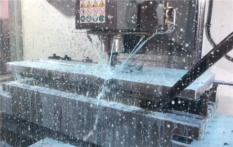 Parts being machined on Midaco Pallet Changer pallet inside VMC with CNC coolant spray on VMC window